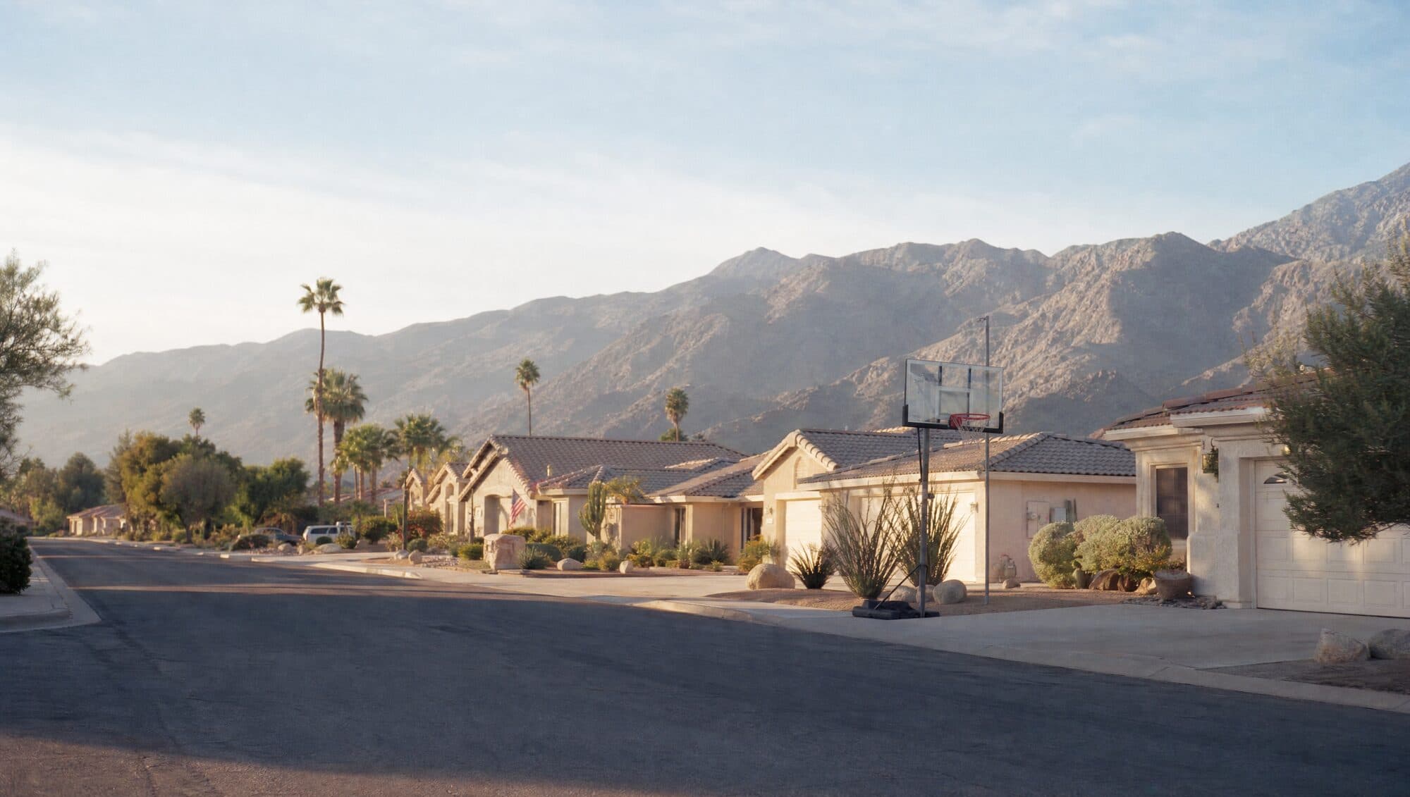 A quiet street of single-story homes with a basketball hoop and mountains behind